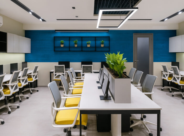 Modern open-plan office with bright overhead lighting, spacious white desks arranged in clusters, ergonomic chairs with yellow seats, and built-in monitors. The space features a striking blue accent wall, stylish hanging light fixture, shelves displaying small green potted plants, minimalist storage cabinets, and a row of tall greenery at the center for a fresh, organized look.