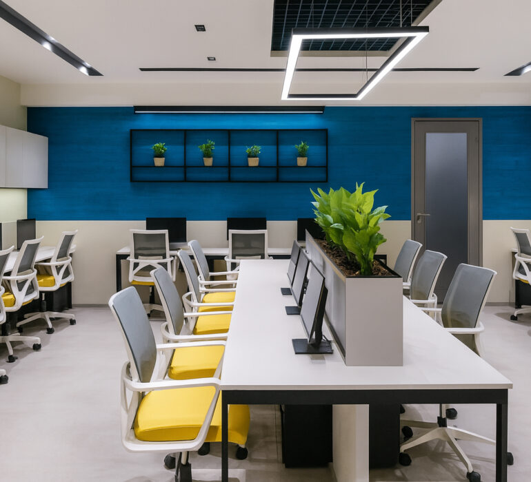 Modern open-plan office with bright overhead lighting, spacious white desks arranged in clusters, ergonomic chairs with yellow seats, and built-in monitors. The space features a striking blue accent wall, stylish hanging light fixture, shelves displaying small green potted plants, minimalist storage cabinets, and a row of tall greenery at the center for a fresh, organized look.