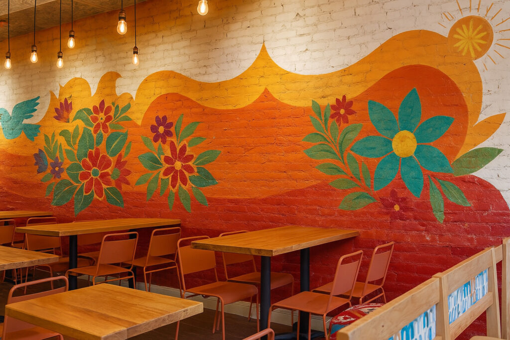 Vibrant café interior with colorful floral mural on brick wall, wooden tables, orange chairs, and warm hanging bulb lighting.